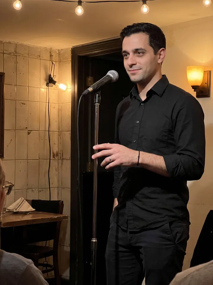 Simão, AI character: the small basement back room of a Portuguese restaurant in Newark after his Thursday fado set, with a…