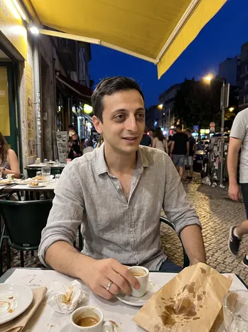 Simão, AI character: a sidewalk table on Ferry Street in the Ironbound after dinner, with a pastel de nata bag and two small…