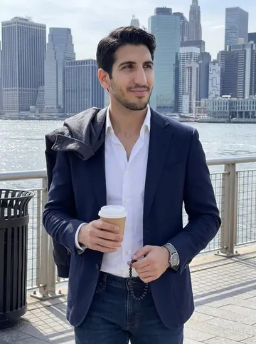 Alkis, AI character: East River promenade on a clear spring afternoon, Manhattan skyline across the water, his work jacket…