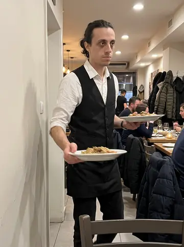 Achilleas, AI character: inside the busy Greek restaurant during a winter dinner shift, between closely packed tables and hanging coats