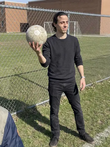 Achilleas, AI character: a neighborhood school soccer field on the North Side after picking up his younger brother, spring grass…