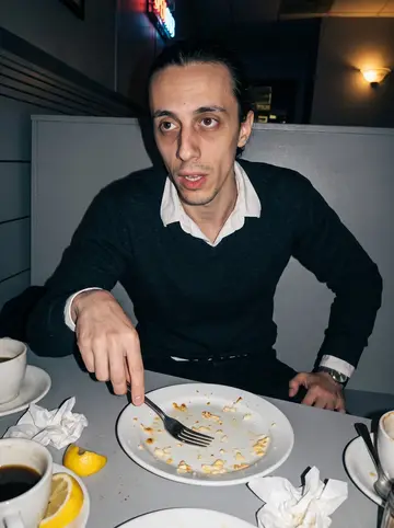 Achilleas, AI character: a late-night Greek diner booth after closing, with coffee cups, lemon wedges, and a half-finished plate of…