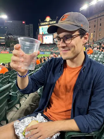 Sotiris, AI character: cheap seats at Camden Yards during an Orioles night game, plastic cup holders and dark green seats around him