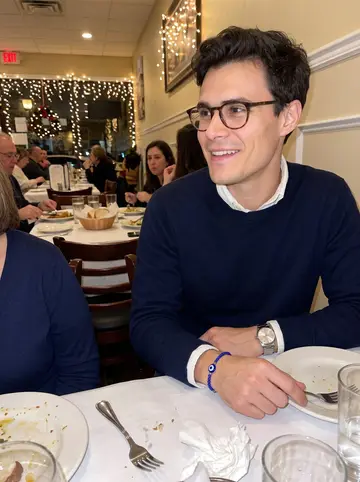 Sotiris, AI character: a crowded Greek restaurant in Baltimore during his yiayia's name day dinner, family shoulders and plates…