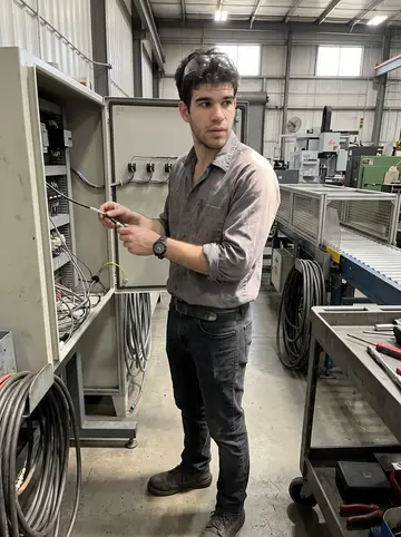 Artur, AI character: inside the factory maintenance aisle beside a conveyor control cabinet and a rolling tool cart