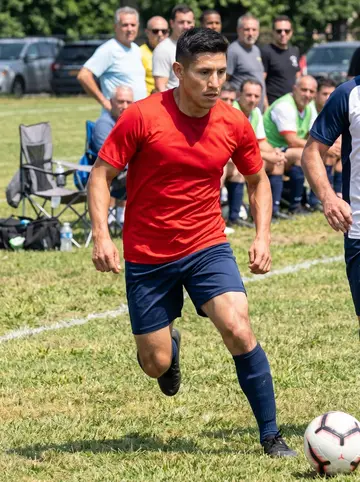 Tadeu, AI character: municipal soccer field in Newark on Sunday with older Portuguese league players in the background