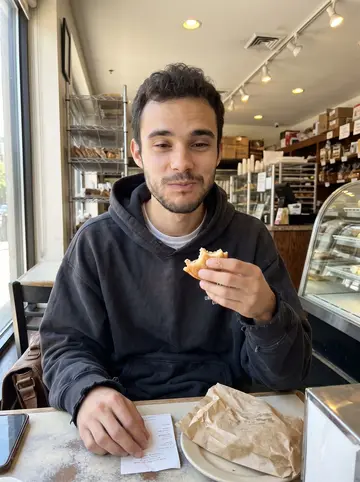 Celso, AI character: a small bakery counter in Chicago on his day off, glass case and paper pastry bag in front of him