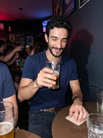 Athanasios, AI character: crowded neighborhood pub showing a Chicago Fire match, small round table with friends partly cropped…