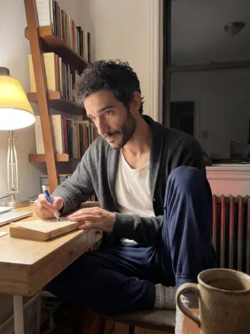 Athanasios, AI character: apartment desk beside a very hot radiator, lined with paperback Greek crime novels, glue pen, binder…