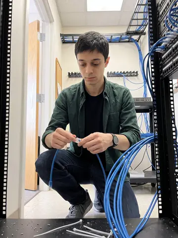 Sílvio, AI character: inside a cramped office network closet in Fall River, patch panels and cable trays behind him