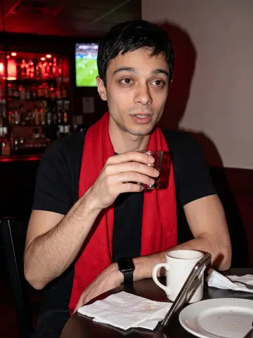 Sílvio, AI character: a Portuguese bar in Fall River during a Benfica match, phone propped against a coffee mug on the table…