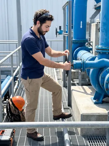 Pavlos, AI character: a Tampa water treatment plant catwalk beside blue pipes and a pump housing during a clear weekday shift