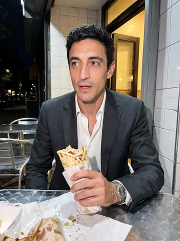 Takis, AI character: outside a small souvlaki place in Astoria at night, tiled wall and metal outdoor table behind him