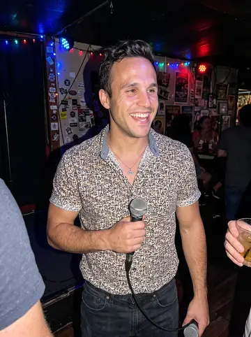 Georgios, AI character: a small Tampa dive bar on karaoke Thursday, stage corner with colored lights and friends cropped at the edges