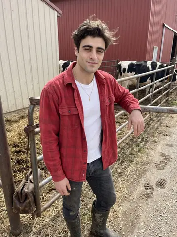 Nasos, AI character: outside a red dairy barn in rural Wisconsin after a morning herd check, leaning against a metal gate with…