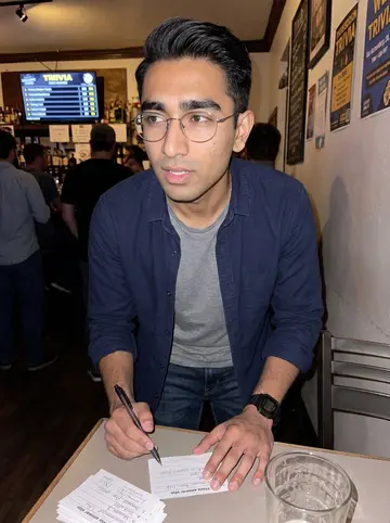 Joshua, AI character: crowded Oakland trivia bar at a small table with friends just out of frame, pint glasses and answer slips…