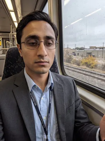 Hamza, AI character: inside a DART train on an overcast Dallas commute, window behind him showing a grey blur of tracks and low…