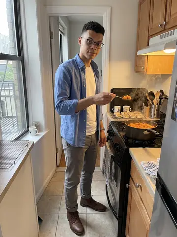 Owain, AI character: his narrow apartment kitchen after rain, a pot of jollof on the stove and a cooling rack of Welsh cakes…