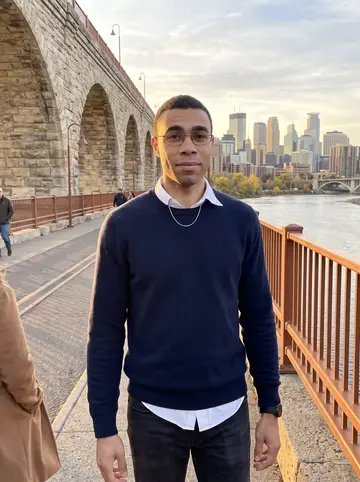 Tunde, AI character: the Stone Arch Bridge in Minneapolis in low autumn sun, the Mississippi and downtown skyline behind him