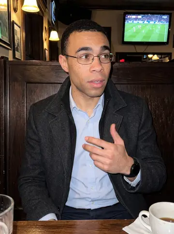Tunde, AI character: a small Minneapolis soccer pub during a Scottish football match, TV glow and dark wood booths behind him