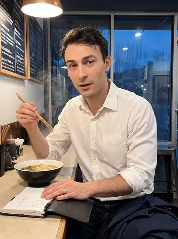 Kunal, AI character: a narrow Denver noodle bar on a rainy evening, seated alone at the counter with steam rising from a bowl…