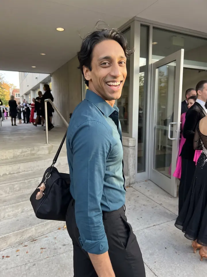 Raymond, AI character: outside a community ballroom competition venue after afternoon practice, glass doors and a few dressed-up…