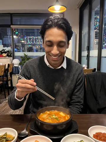 Raymond, AI character: small Korean restaurant on a cold evening, bowl of kimchi stew and side dishes on the table, holiday…