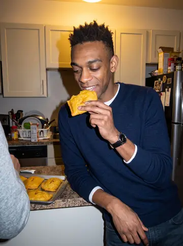 Jelani, AI character: a warm apartment kitchen in Minneapolis during a small family dinner, trays of Jamaican patties cooling on…