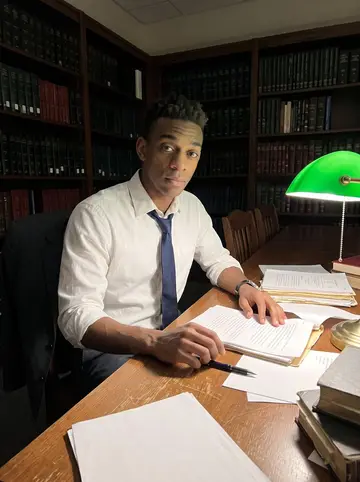 Jelani, AI character: a nearly empty law library reading room after hours, stacks of old courtroom transcripts and a worn…
