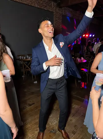 Tyrone, AI character: a crowded wedding reception in a Manayunk event hall, dance floor lights, plastic cups on cocktail tables,…