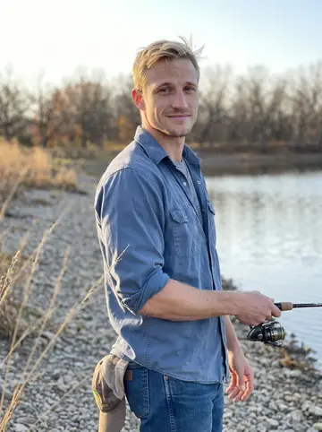 Hank, AI character: the gravel bank of Lake Carl Blackwell just after sunrise, still water and bare trees behind him