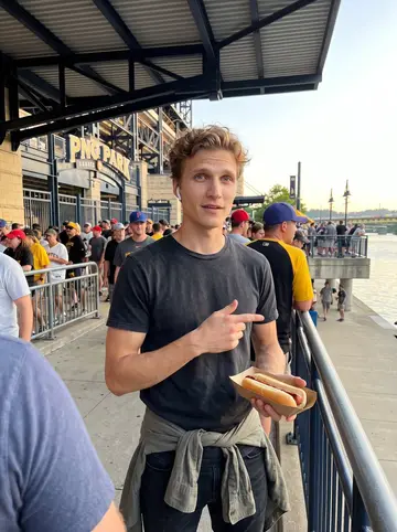 Oscar, AI character: outside PNC Park before a Pirates game, standing near the riverwalk with the stadium crowd behind him