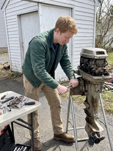 Dominick, AI character: a driveway beside a small garage in South Buffalo, an old outboard boat motor clamped to a stand with…