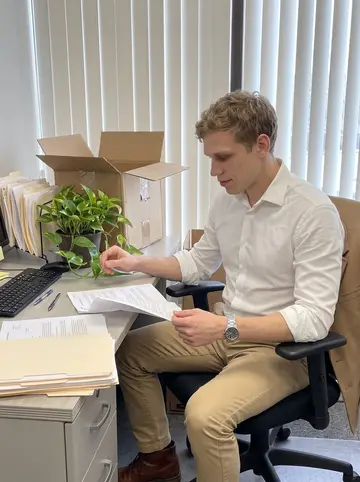 Joey, AI character: his small new insurance office in Columbus, with a tidy desk, a pothos plant named Gary, paper folders,…