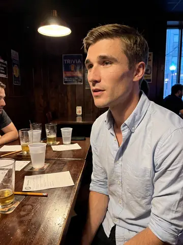 Sebastian, AI character: a narrow Philadelphia trivia bar with sticky wooden tables and a chalkboard menu blurred behind him