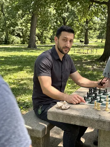 Muhammad, AI character: a concrete chess table in Tower Grove Park, seated across from an older man just out of frame, trees and…