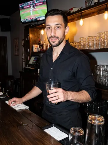 Muhammad, AI character: inside the Cherokee Street bar after last call, with stacked glasses, dark wood, and a low Cardinals game…