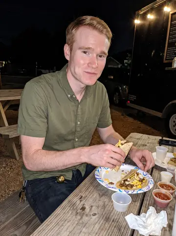 Myles, AI character: outside a taco trailer on South Congress at night, picnic table, paper plate of breakfast tacos and a…