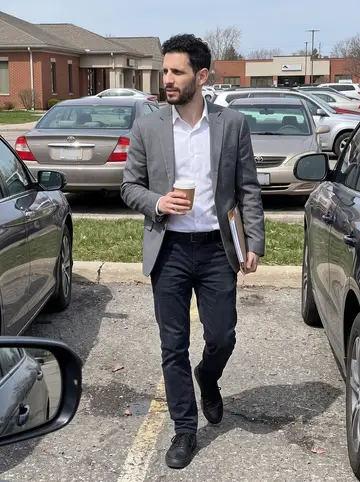 Kareem, AI character: parking lot outside a Dearborn insurance office after a client meeting, his used sedan and a row of low…