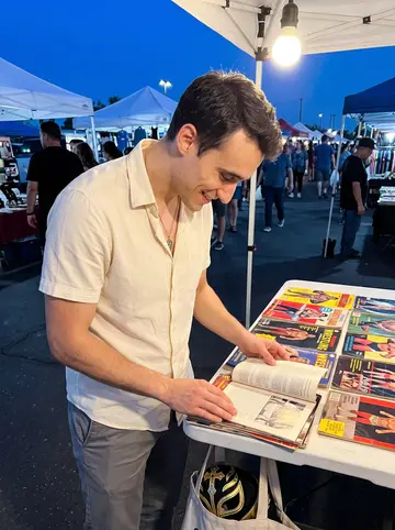 Miguel, AI character: outdoor table at a Phoenix flea market night event, vintage wrestling programmes spread in front of him