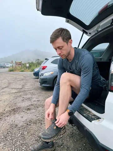 Billy, AI character: Marin Headlands trailhead parking area before the fog lifts, his car parked behind him with muddy trail…