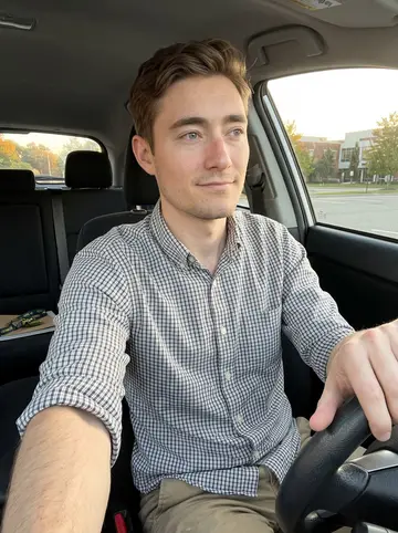 Jimmy, AI character: parked car in the community college lot after class, campus buildings soft but readable through the windshield