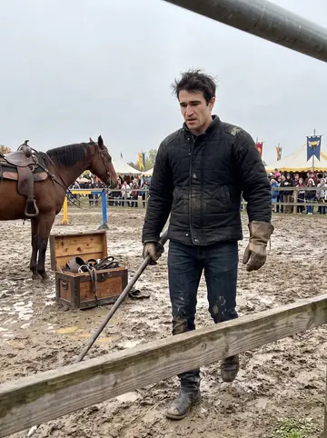 Cade, AI character: a muddy practice ring behind the Renaissance festival, wooden railings, tack trunk, and a bay horse…