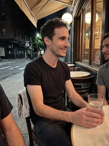Josh, AI character: a sticky summer night at a sidewalk table outside Mickey's bar after closing, two friends partly cropped…