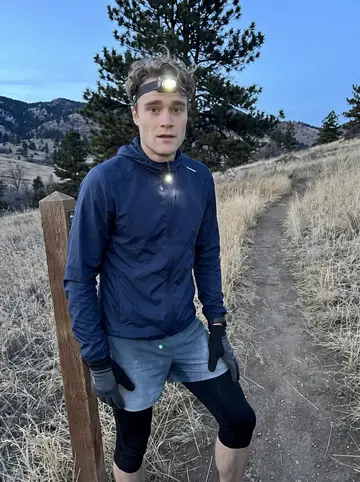 Drew, AI character: a foothills trail above Boulder just after dusk, dry grass and dark pines behind him, headlamp strap…