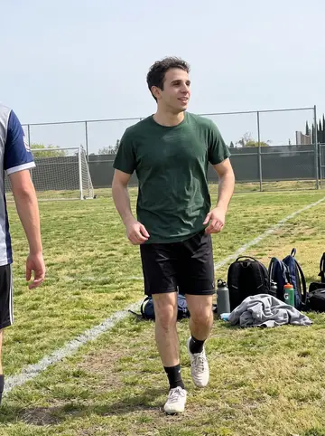 Enrique, AI character: a neighborhood soccer field in Sacramento on a Sunday morning, patchy grass and backpacks along the sideline
