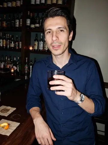 Kristian, AI character: a narrow Baltimore cocktail bar after a Friday practice night, dark wood counter and shelves behind him