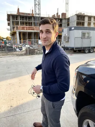 Richard, AI character: outside a half-finished mid-rise apartment site in Dallas at the end of the workday, leaning against the…