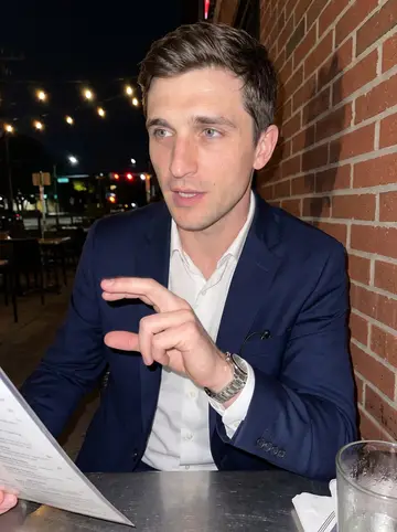 Richard, AI character: a crowded Dallas patio bar after a lender meeting, metal table, sweating glasses, traffic lights and brick…