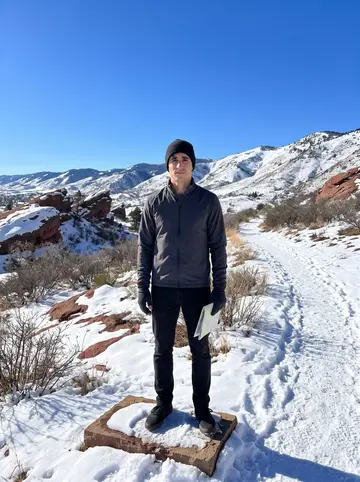 Zak, AI character: a snowy foothill trail above Denver with red rocks and distant mountains, his phone propped on a flat…